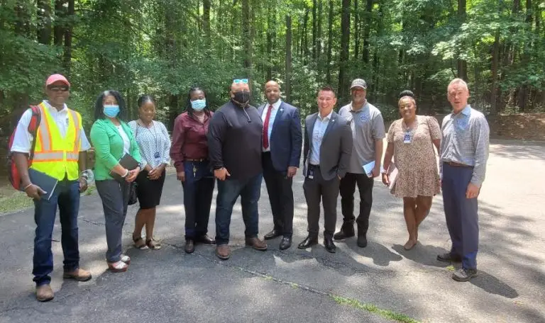 A group of people standing on a paved path in a forested area, some wearing masks and formal clothing.