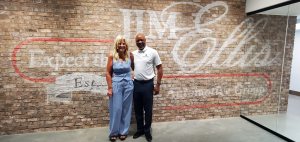 Two people stand smiling in front of a brick wall with Jim Ellis and other text painted on it.