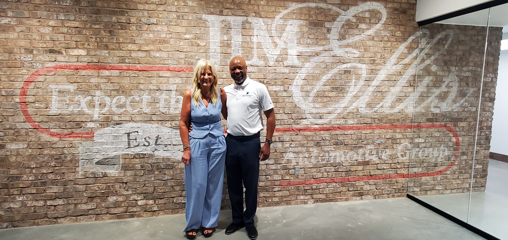 Two people stand smiling in front of a brick wall with Jim Ellis and other text painted on it.