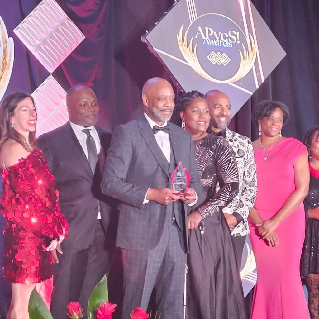 A group of people in formal attire pose with an award at the APyeS! Awards in front of a decorated stage.