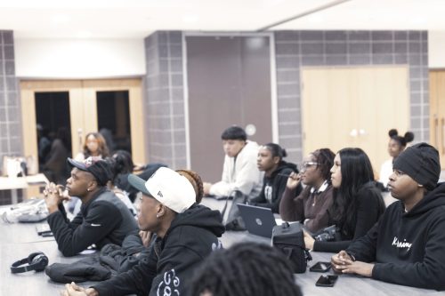A group of students sit at tables, attentively listening in a modern classroom setting.
