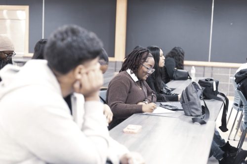 Students seated at desks in a classroom, with one student writing and others listening attentively.