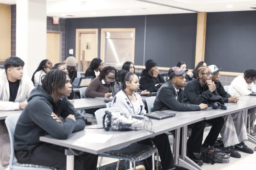 A group of students sit attentively at desks in a classroom, facing forward and listening to a speaker.