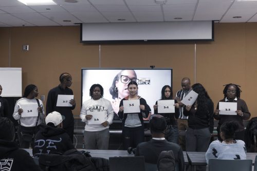 Eight people stand in front of a classroom, holding signs with numbers, as others watch and a video call is on screen.
