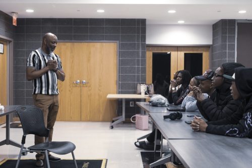 A man stands and speaks to a group of people seated at a long table in a classroom setting.