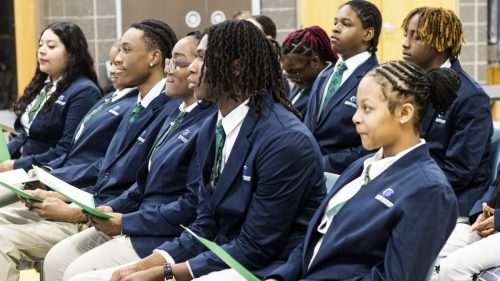 A group of students in blue blazers and green ties sit in rows, holding papers, at an indoor event.