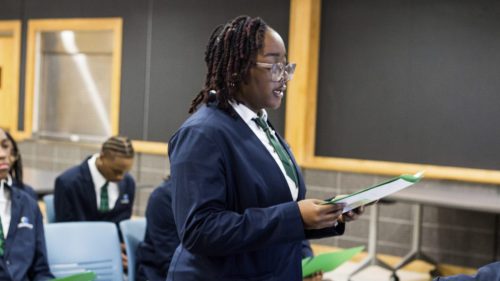 A student in a suit speaks while holding papers in a classroom, with others seated in the background.