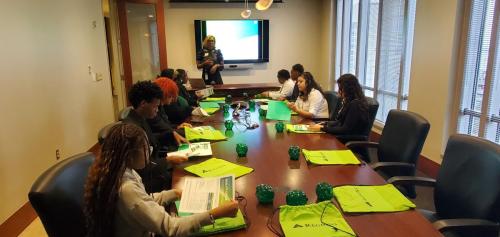 People in a conference room sit at a table, engaging in a financial literacy presentation with green folders before them.