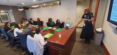 A woman presents on financial literacy to a group seated around a conference table with green folders and water bottles.
