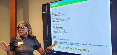 Woman presenting on financial literacy, gestures near a screen displaying a persona profile and financial scenario.