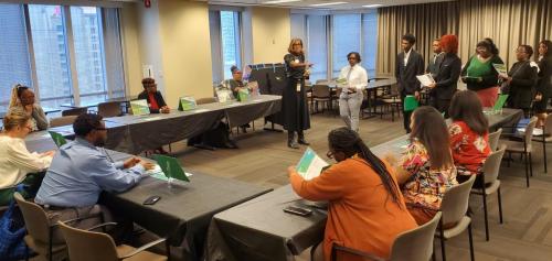 A group in a meeting room discusses financial literacy, with a woman standing and speaking while others take notes.