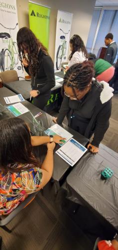 People engaging at a job fair booth, discussing brochures on financial literacy with a banner in the background.