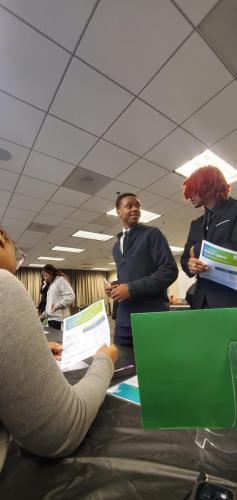 People in formal attire discuss financial literacy at a table, holding papers in a well-lit room with a tiled ceiling.