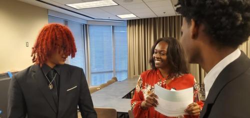 Three people in formal attire discuss financial literacy, smiling in a meeting room. One holds a piece of paper.