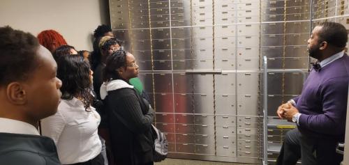 A group learns about financial literacy from a speaker inside a vault lined with safety deposit boxes.