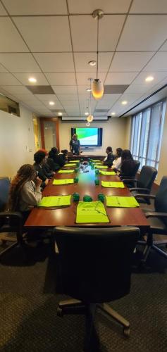 Meeting room with people seated at a long table, presenter discussing financial literacy by a screen displaying a presentation.