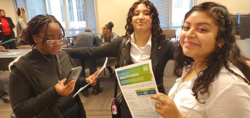 Three people smiling and holding papers on financial literacy, with others in the background.