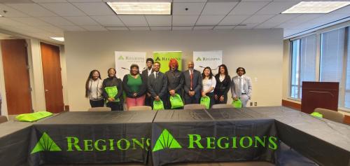 A group stands behind a Regions-branded table with green bags, promoting financial literacy in a conference room.
