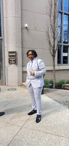 Person in a light gray suit smiles, standing by a building with Salesloft, Holland  Knight, and financial literacy signs.