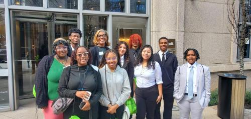 A group of ten people smiles in front of a building entrance, dressed in business attire, celebrating financial literacy with green bags.
