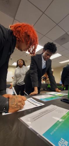People in business attire signing forms at a table while a woman observes, promoting financial literacy in a tiled-ceiling room.