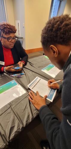 A woman with glasses sits at a table, discussing financial literacy with a person writing on a form.