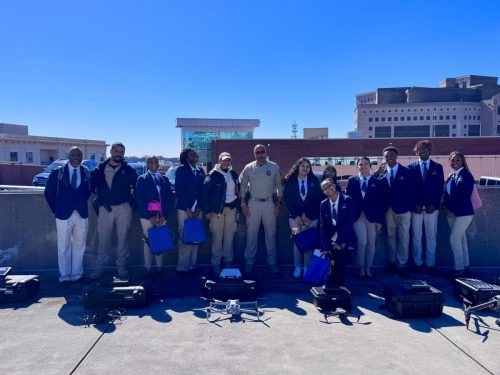 A group of people stand outdoors in formal and uniform attire with drones and equipment in front of them.