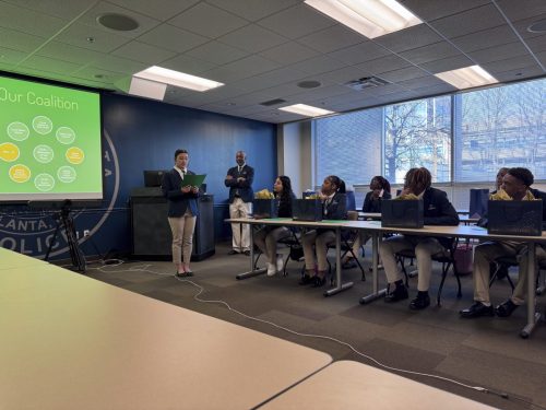 A presenter speaks to a group of students seated at tables, with a coalition chart displayed on a screen.