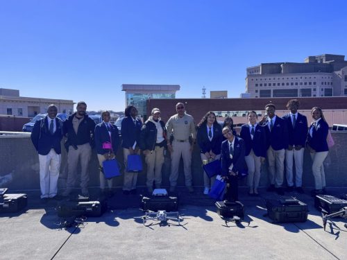 A group of people in uniforms and suits standing outside with drone equipment on the ground in front of them.