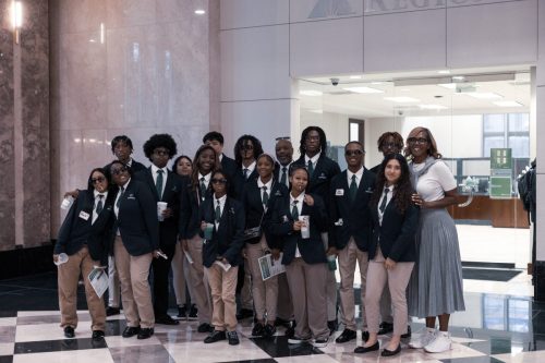 A group of students in uniforms pose together indoors with an adult woman, all smiling at the camera.