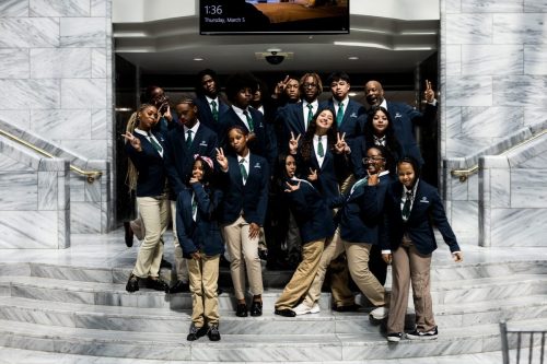 A group of students in blue blazers pose together on marble stairs, making peace signs and smiling.