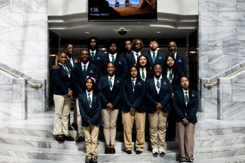 A group of students and adults in navy blazers and khakis pose together on marble stairs indoors.