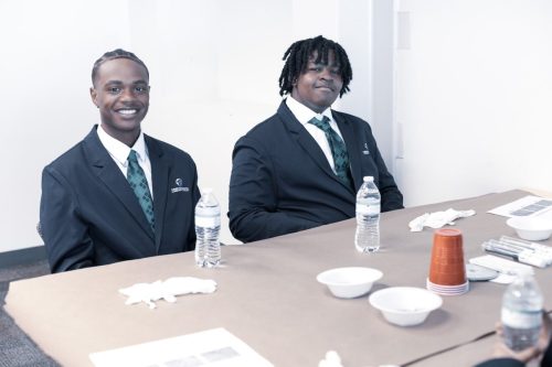 Two young men in suits sit at a table with water bottles, cups, and papers, smiling at the camera.