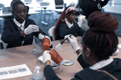 Three students in gloves and suits examine objects and brush powder at a classroom table during a forensic activity.