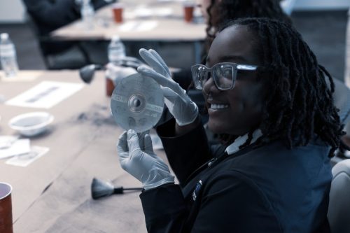 Person wearing gloves and glasses smiles while holding up a CD at a table with forensic investigation tools.