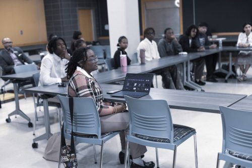 Students sit at tables in a classroom, some with laptops open, attentively listening to a presentation.