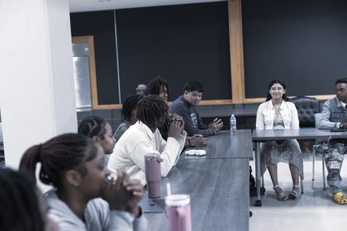 A group of people sit around tables in discussion in a classroom or meeting room setting.