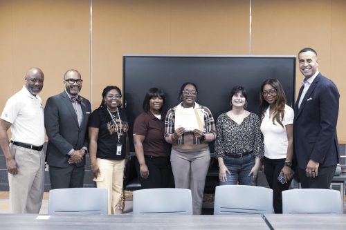 A group of eight people smiling, one person in the center holding a certificate in front of a large screen.