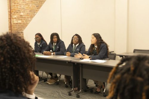 Four people in business attire sit at a conference table, speaking to an audience in a meeting room.