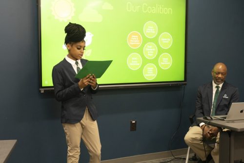 A young person presents with notes beside a seated man and a green coalition chart on a screen.