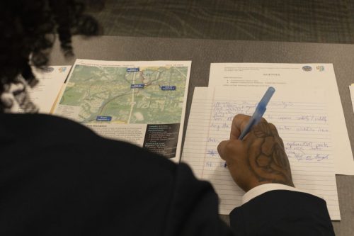 Person takes notes beside a printed map and agenda at a meeting, showing a tattooed hand holding a pen.