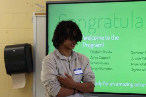 Person with long hair stands in front of a presentation screen displaying welcome message and names.