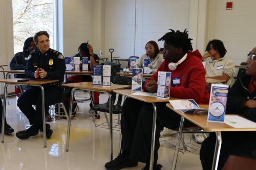 A police officer talks to students seated at desks in a classroom setting.